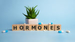 Block letters spell the word "hormones" in front of a plant and blue background. Various pills are scattered around the photo.