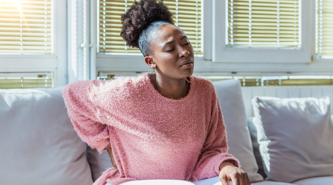 A young Black woman sits on a couch, with a hand on her lower back where she is experiencing pain related to her uterine fibroids