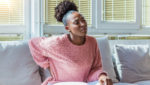 A young Black woman sits on a couch, with a hand on her lower back where she is experiencing pain related to her uterine fibroids