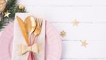 A pink and gols ilverware set sits on top of a pink dinner plate. The background is decorated for Christmas.