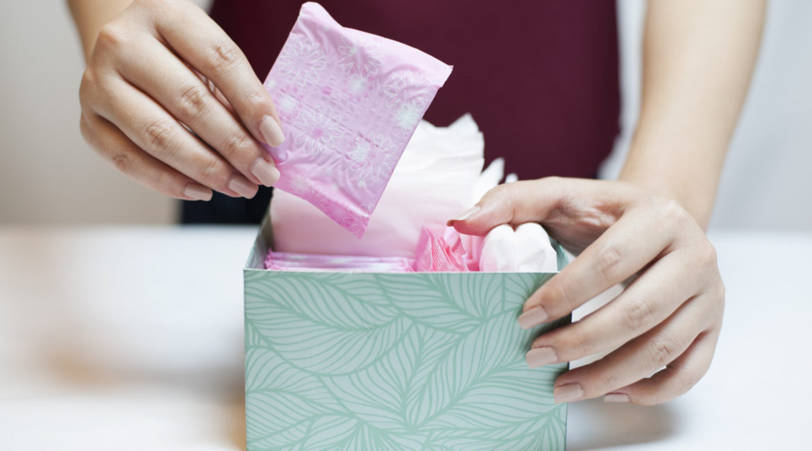 A woman reaches into a box to pull out a menstrual pad wrapped in pink plastic.